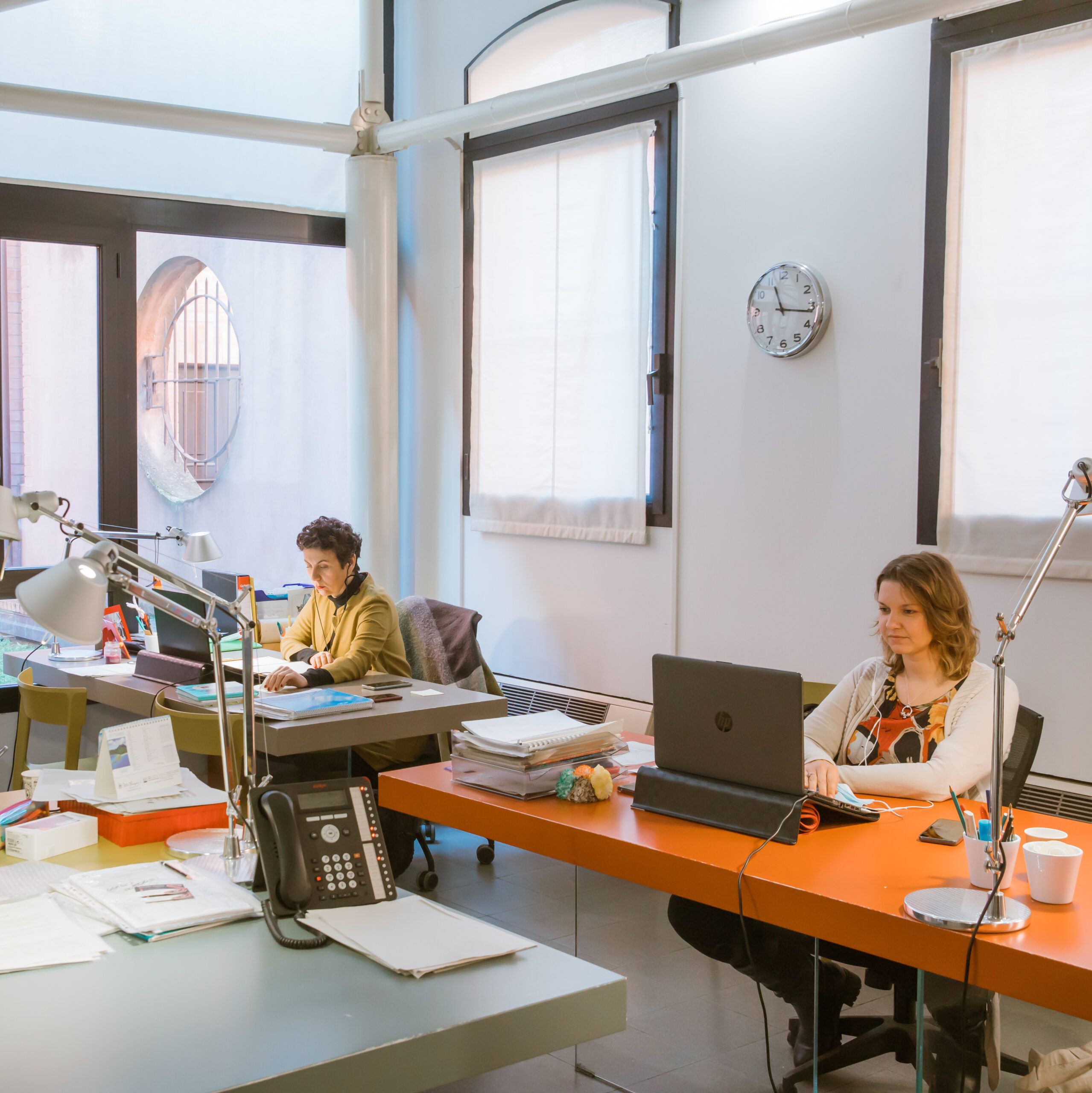 Due donne lavorano al pcl, ognuna all propria scrivania. L'ambiente è quello di un ufficio, c'è un orologio a muro e ci sono delle finestre.