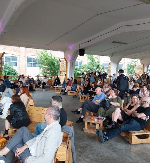 Gruppo numeroso di persone sedute su panche di legno con schienale sotto una grande tettoia ascoltano qualcuno o guardano qualcosa.