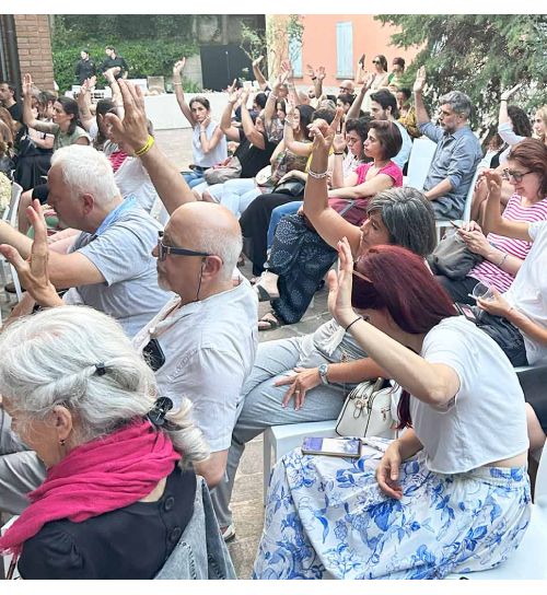 Persone sedute su sedie all'aperto alzano la mano per votare. La scena è molto luminosa.