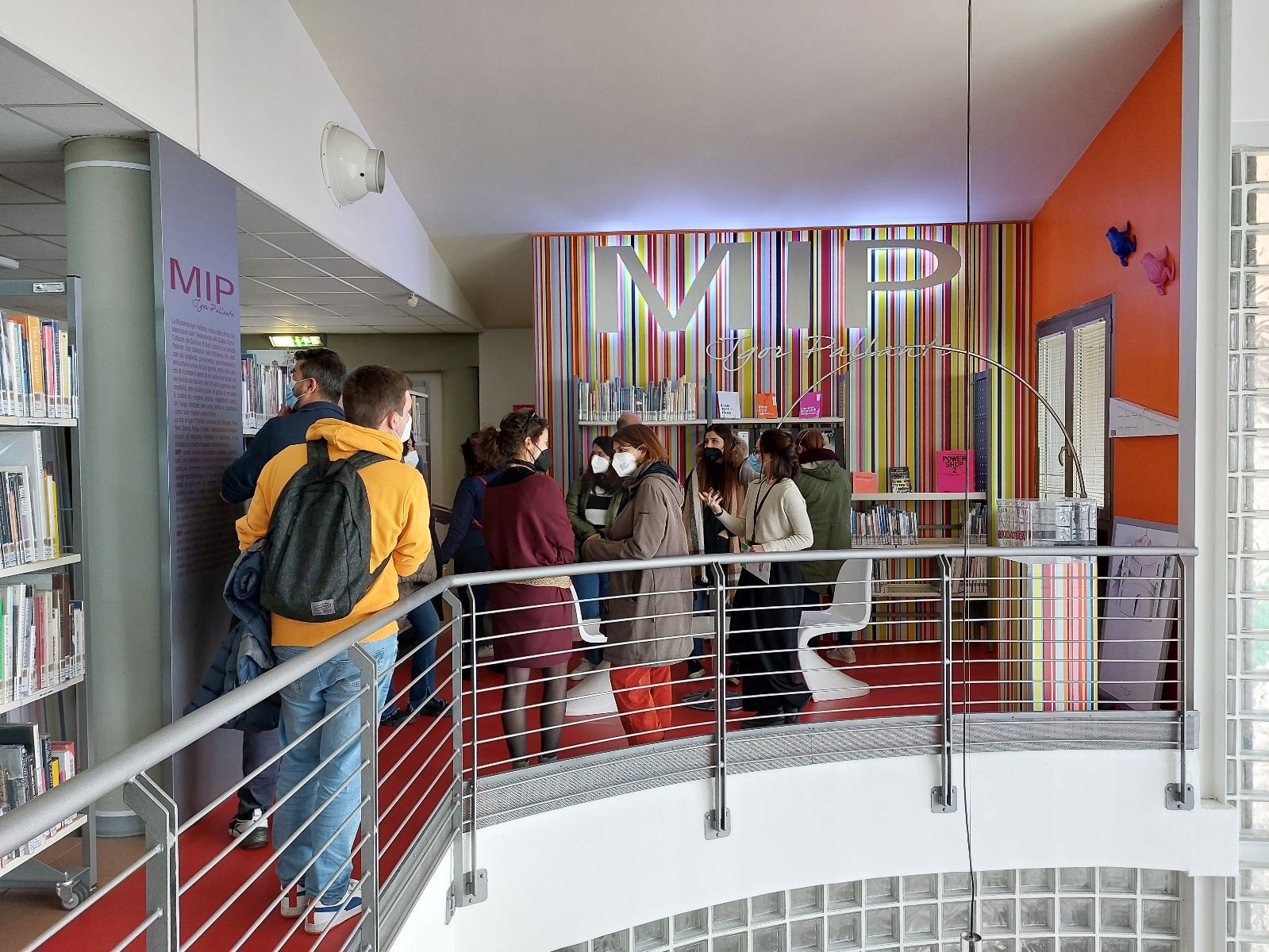 Gruppo di persone in una biblioteca. Lo spazio ha le pareti rosse e si trova al secondo piano, si può vedere una ringhiera di metallo bianca che circonda le persone e la scritta "MIP" sullo sfondo.