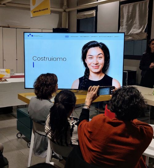 Foto di uno schermo durante la presentazione di Oplà. Sullo schermo il viso di una ragazza sorridente con a fianco la scritta "costruiamo". Una donna, girata di spalle, sta scattando una foto allo schermo.