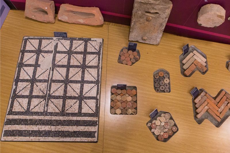 Immagine ingrandita di esposizione museale di mosaico rappresentate le domus romane al museo di Claterna. Di fianco tessere mosaico policrome in pietra. Il tutto ha didascalie per il pubblico