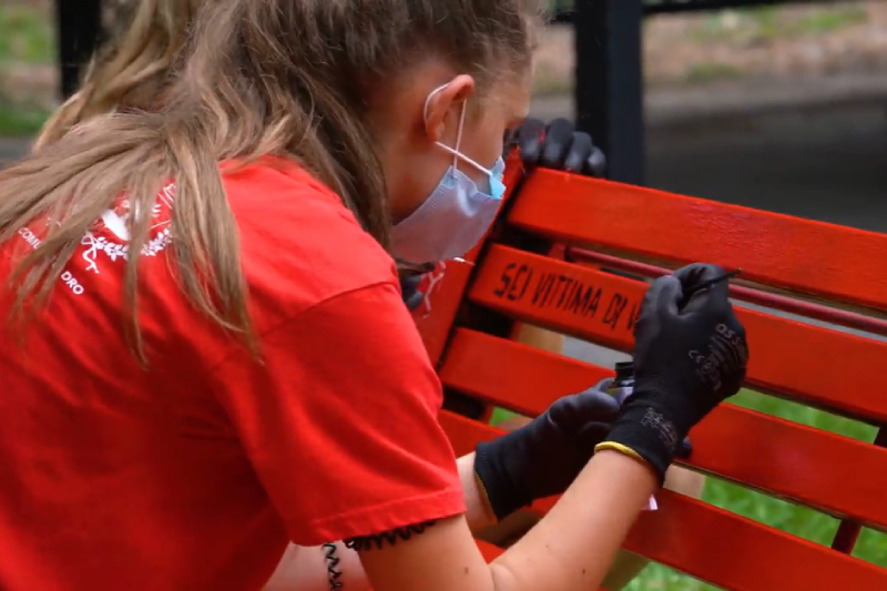 Bambina con maglia rossa, mascherina e guanti neri dipinge una panchina con un pennello