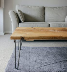 In primo piano un tavolino moderno effetto legno, e sotto a questo un tappeto a pelo morbido grigio. Sullo sfondo un divano dal design contemporaneo, grigio chiaro.