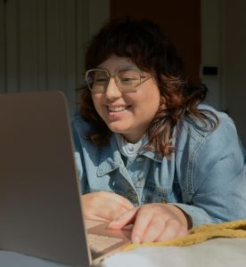 Ragazza giovane sorride guardando lo schermo di un computer. Indossa gli occhiali, ha i capelli lunghi fino alle spalle e porta una camicia di jeans chiara.