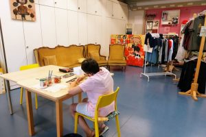Persona che frequenta l'atelier artistico protetto Marakanda è fotografata di schiena mentre è seduta su una sedia gialla. La donna sta disegnando. Sullo sfondo alcuni dei prodotti realizzati dal laboratorio.