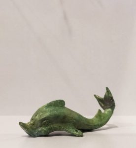 Piccola statuetta verde ritraente il delfino sacro del Dio Apollo, appoggiata su un tavolo bianco. La figura è leggermente ondulata con la coda che punta verso l'alto.