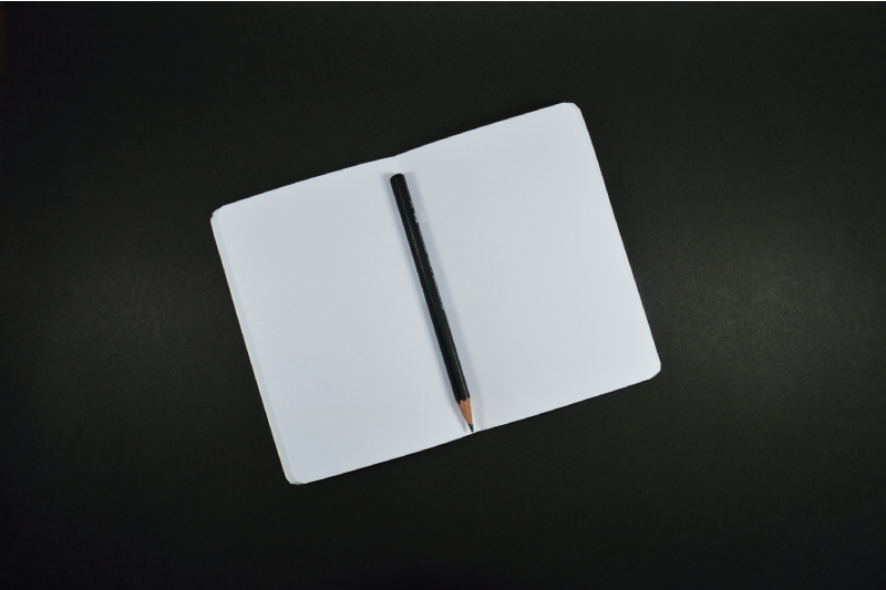 Matita nera appuntita posta a metà di un quaderno bianco, vuoto, tutto su sfondo nero. Il quaderno non ha nè righe nè quadretti ed è aperto completamente. 