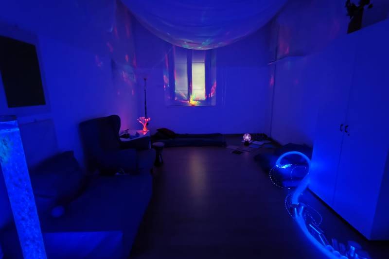Foto della stanza multisensorisale nel centro di Borgonuovo. La stanza è buia, illuminata da luci secondarie blu e rosse provenienti da lampade e proiettori. Al centro è vuota e ai lati sono presenti lampade e divani.