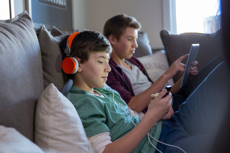 Due adolescenti giocano con dispositivi digitali. Sono posti di profilo, quello più vicino all'inquadratura è appoggiato su dei cuscini bianchi e grifi indossa una maglietta verde e guarda lo smartphone con delle cuffie rosse. Quello più sullo sfondo ha in mano un ipad 