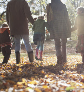 Gruppo di adulti, adulte, bambine e bambini si tengono per mano. Sono fotografati di spalle e camminano su una distesa di foglie cadute dai colori autunnali. L'atmosfera è luminosa, calda, di festa.