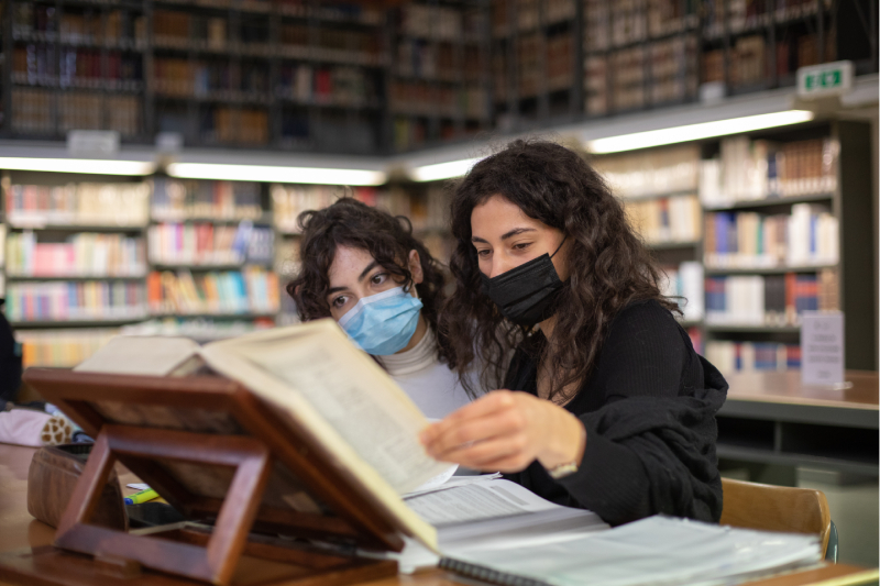 Due ragazze indossano una mascherina e sfogliano un libro all'interno di una biblioteca.