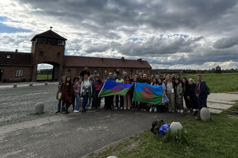 Un gruppo di ragazzi e ragazze del progetto TRACER in visita al campo di concentramento di Auschwitz. Il gruppo espone la bandiera internazionale del popolo rom.