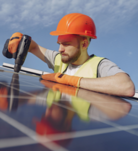 Operaio con casco arancione e giubbotto di sicurezza giallo utilizza un trapano per lavorare su un pannello solare, sotto un cielo sereno