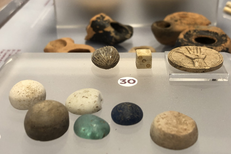 vetrina espositiva del Museo di Claterna con vari oggetti antichi, numerata con il numero "30". Gli oggetti esposti includono pietre di diverse forme e colori, un dado antico con numeri, una placca rotonda con un'incisione a forma di mano. Sullo sfondo, ci sono alcuni pezzi di ceramica o terracotta più grandi e scuri.