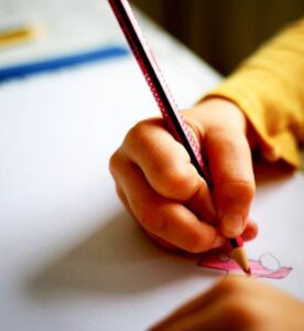 Mano di bambino o bambina che regge una matita colorata con la quale disegna su un foglio bianco. La persona indossa una maglia a maniche lunghe gialla.