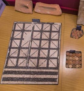 Esposizione su tavolo di resti di mosaico di domus romane al museo di Claterna. Di fianco tessere mosaico policrome in pietra. Il tutto ha didascalie per il pubblico