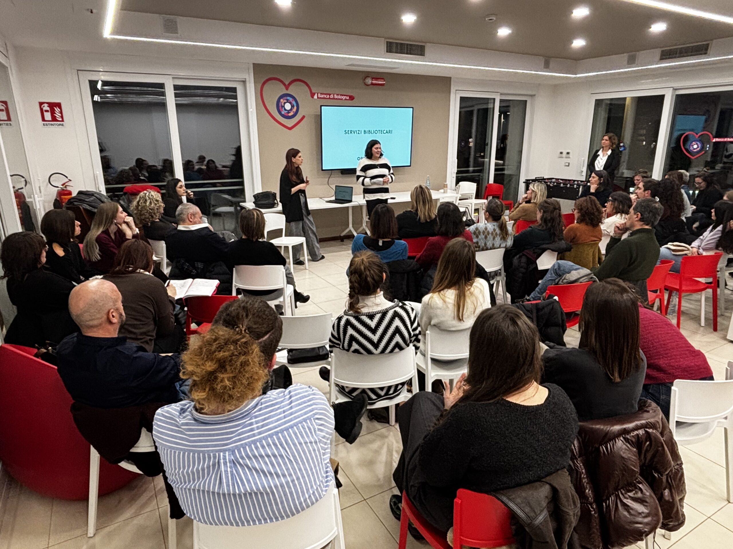 Mini Fuori Orario San Lazzaro, due bibliotecarie parlano alla platea proiettando slides in un televisore