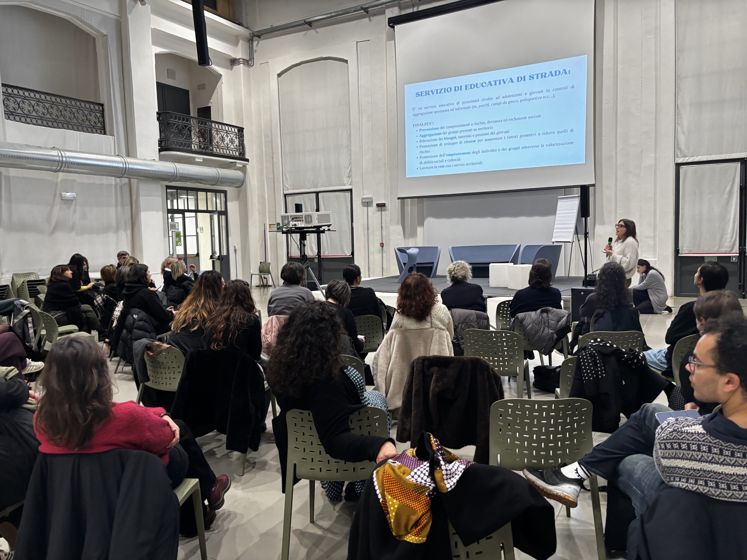 Ragazza parla a paltea in sala molto grande e illuminata con dietro megaschermo e slide con scritto educativa di strada